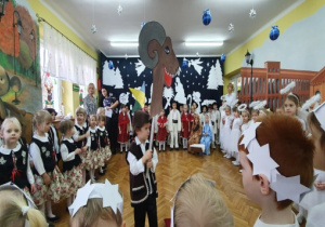 Na środku sali na tle dekoracji świątecznej stoi chłopiec z turoniem, po bokach stoją przedszkolaki.
